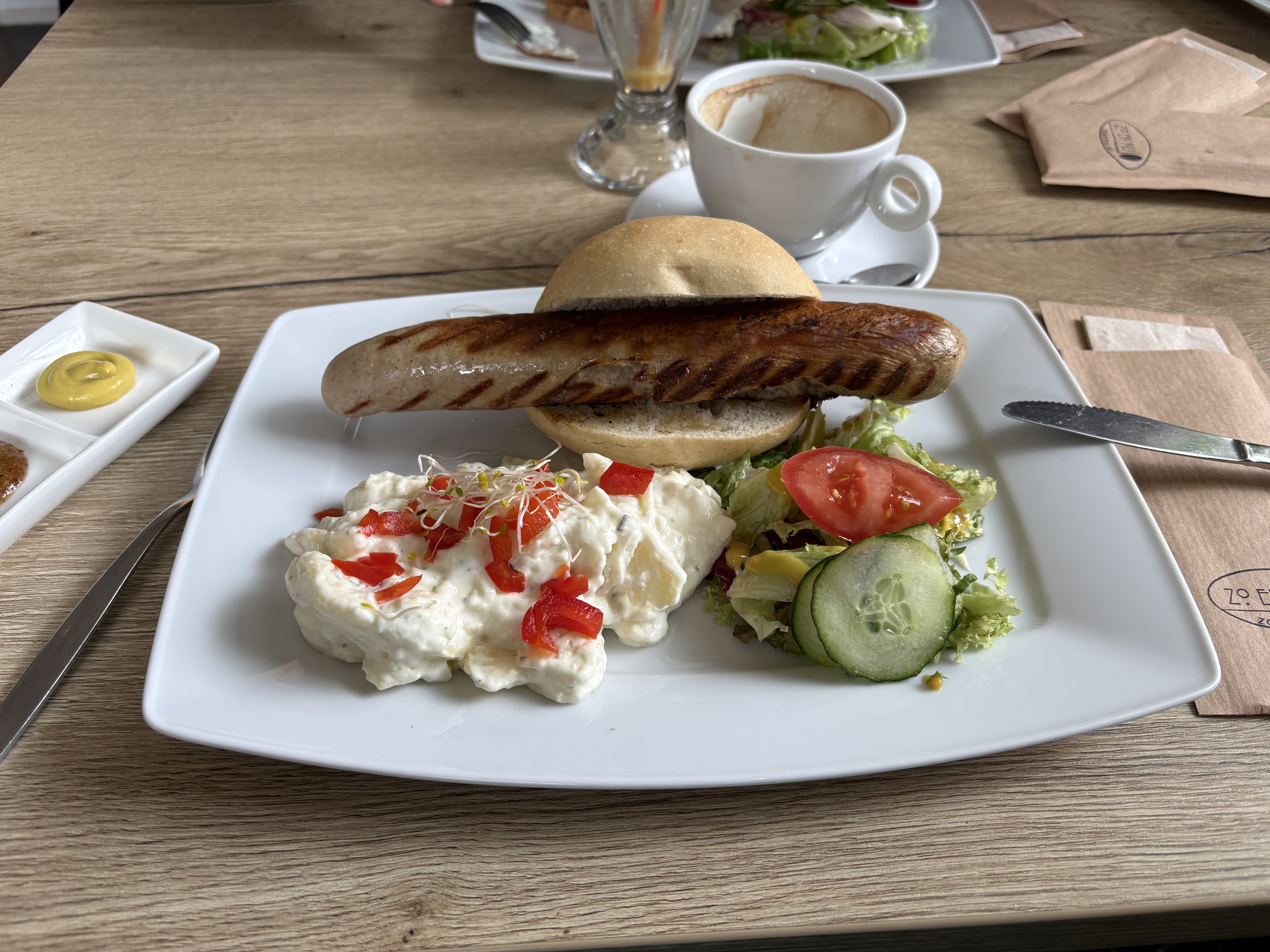 A square plate on a restaurant table filled with a salad, a kartoffel salad and a small white bun with a huge German bratwurst on it.